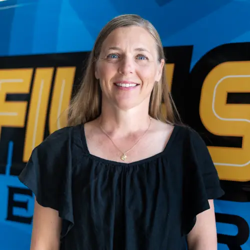 A woman with long hair is standing in front of a blue and yellow wall featuring bold lettering, wearing a black top with ruffled sleeves.